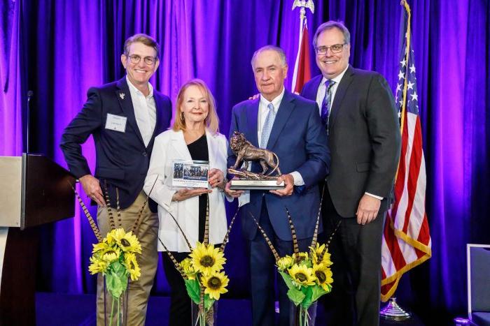 从左到右依次为. Rudin, Senator Ted Lyon, Donna Lyon and Dr. Kelly Reyna stand on stage in front of a purple curtain and the U.S. 和德州旗帜. The Lyons are holding a lion statue. 在人们面前，地板上放着三束向日葵.