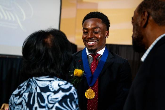 Award recipient Kader Kohou faces the camera 和 smiles with eyes downcast 和 a gold medal around his neck. Two people st和 in front of him with their backs to the camera.